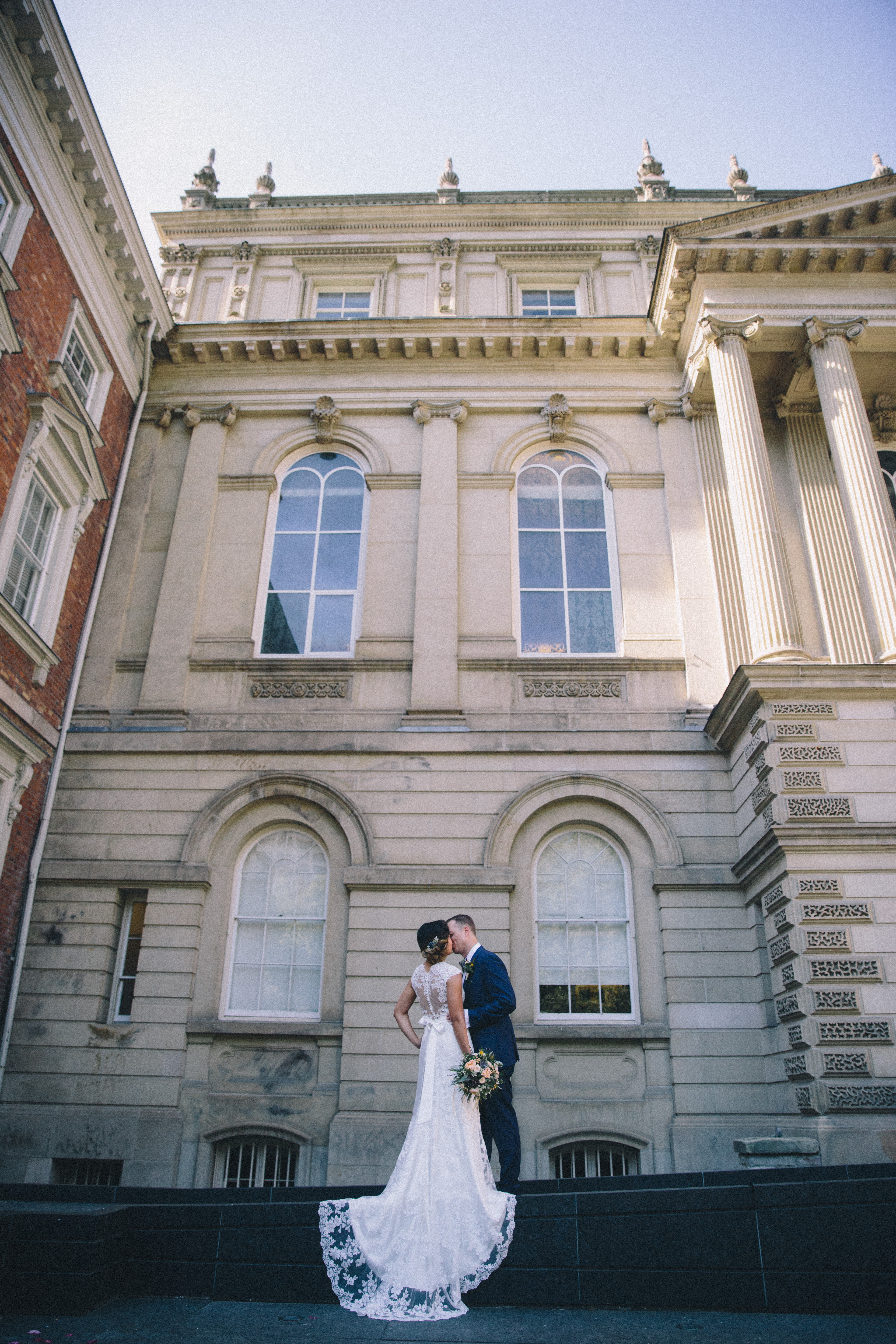 Bride wearing slim fit lace bridal gown with train and bow kisses her groom at Osgoode Hall