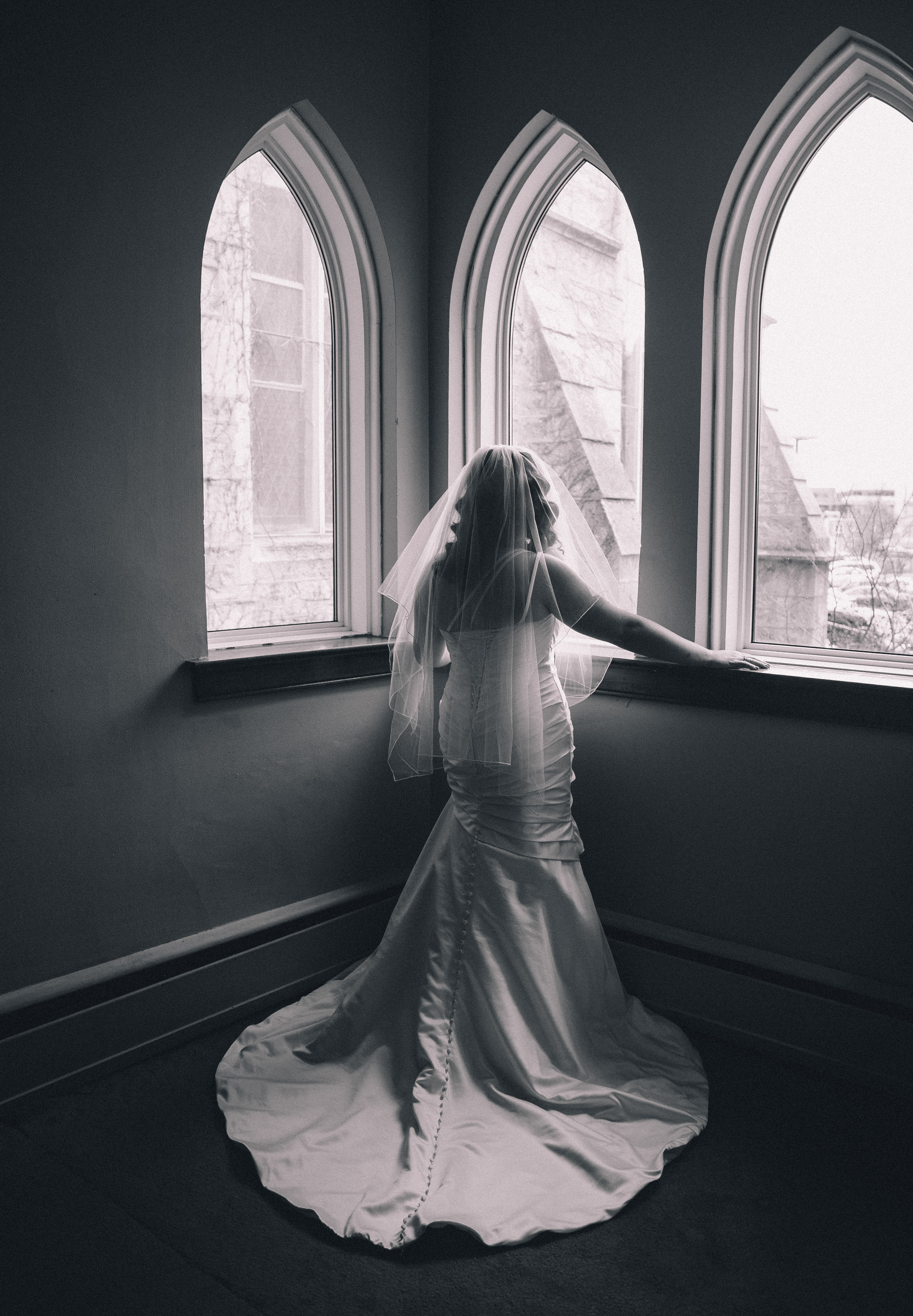 Bride wearing a pleated and gathered mermaid style wedding gown stands looking out the window at Royal City Church in Guelph