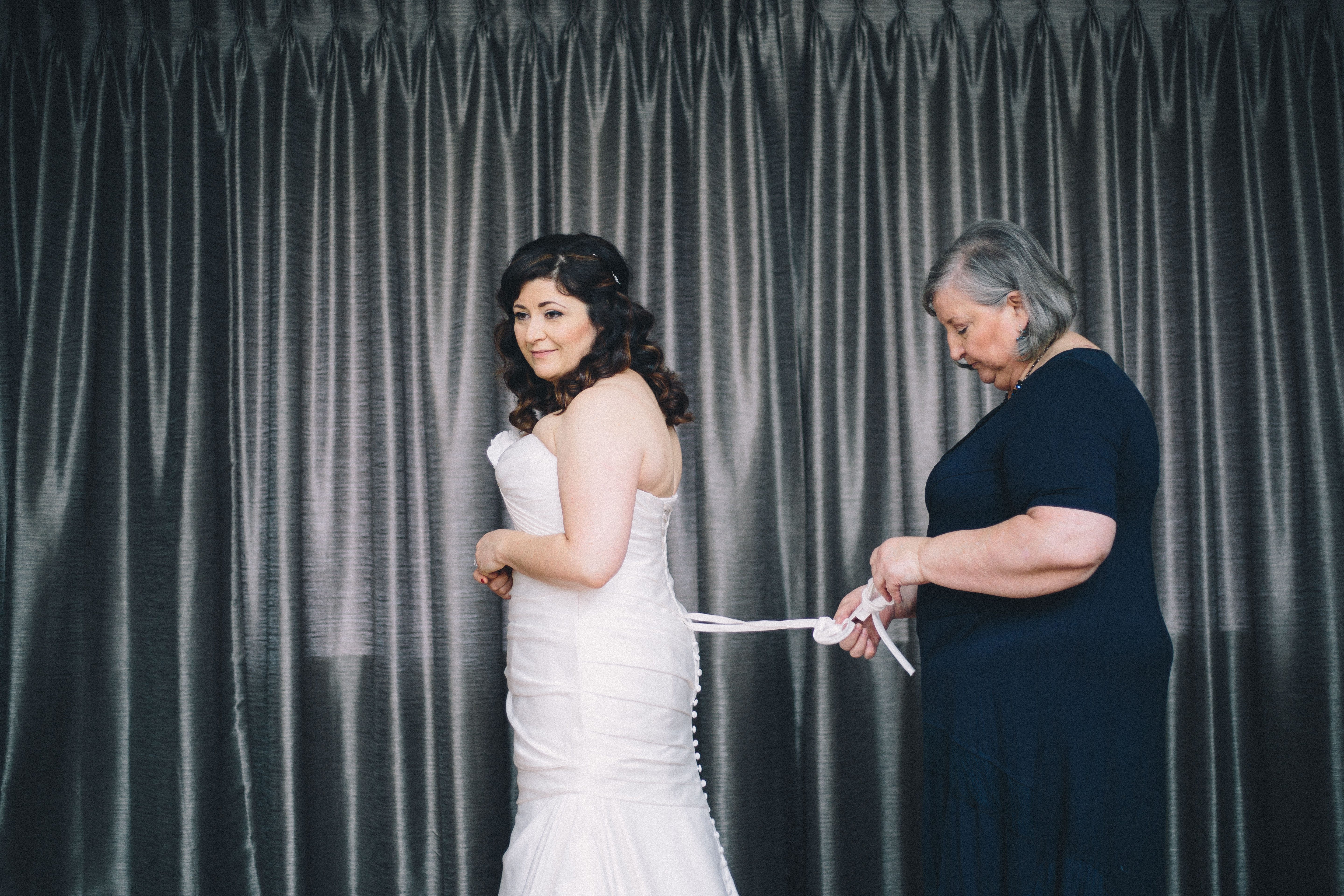 Brunette bride stands holding hands in front of curtains as her Mother lacing up her gathered mermaid style wedding gown