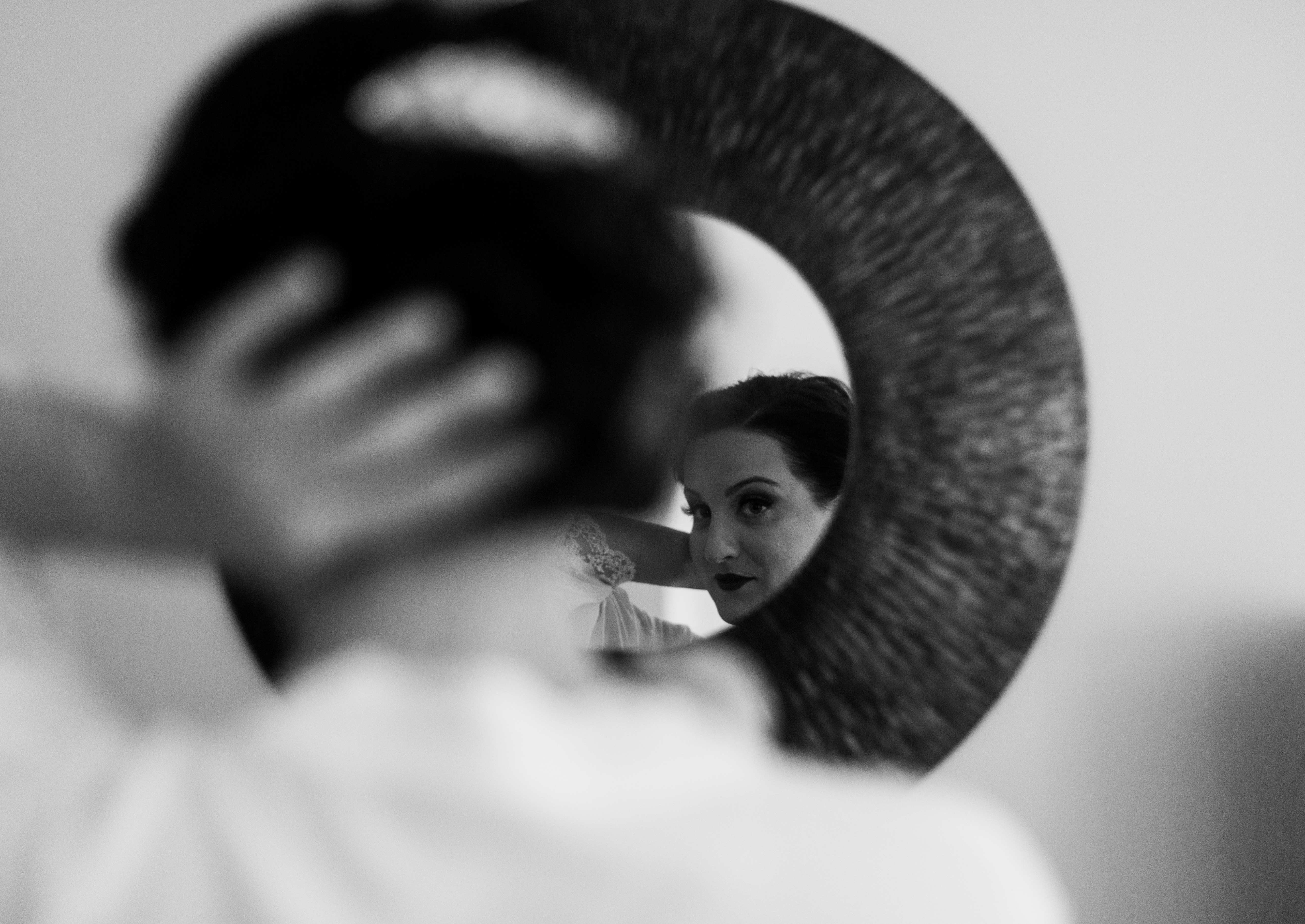 Bride with dark hair and beaded hairpiece looks at her reflection in a rounded mirror before her summer vineyard wedding