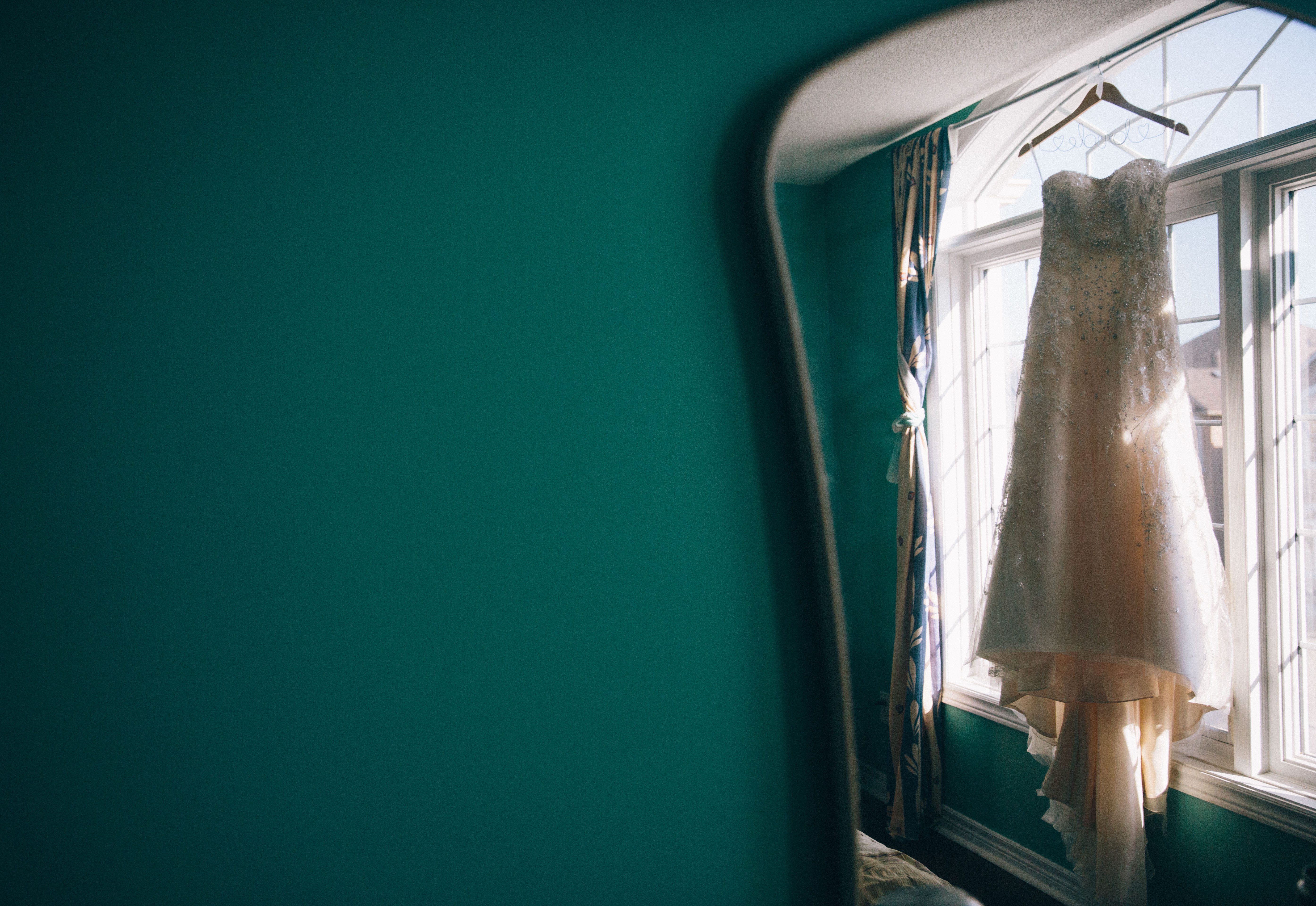 Reflection of beaded sweetheart neckline wedding dress hanging in front of the window next to a teal wall