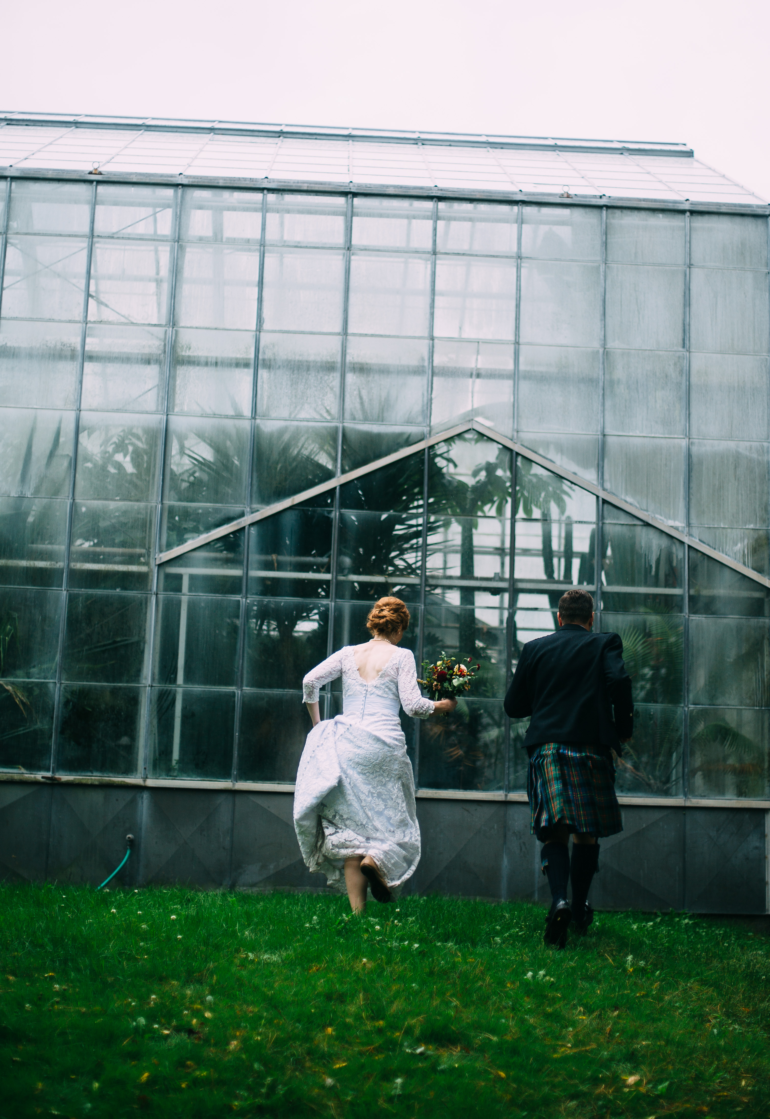 Bride with red hair holds her lace long sleeve wedding gown and runs next to her groom wearing a kilt towards McMaster Biology greenhouse