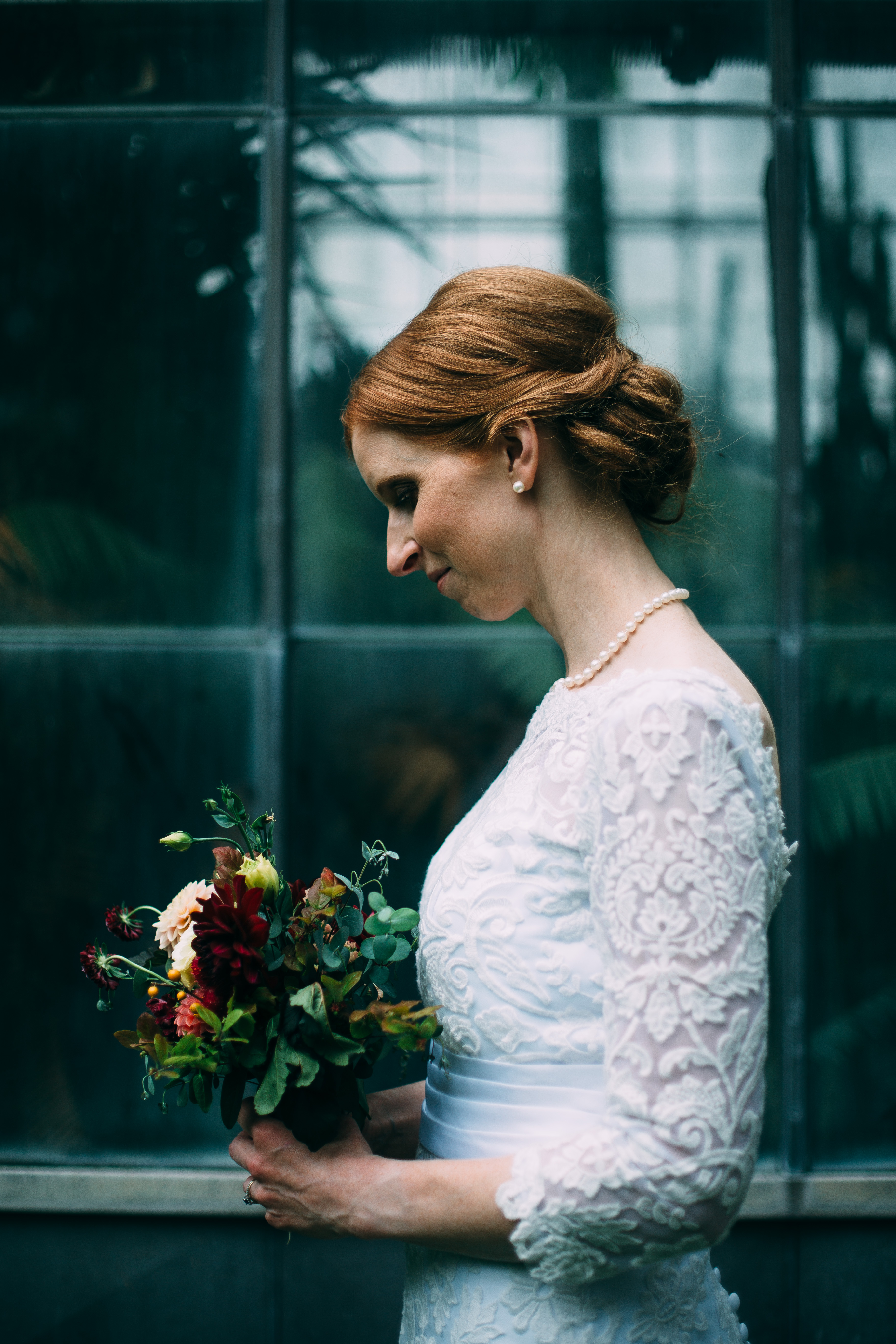 Bride with red hair in chic low bun stands profile against the McMaster University Biology greenhouse and looks down at her loose bridal bouquet of fall florals and foliage